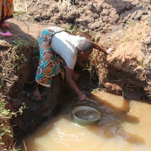 woman fetching water from dirty water hole in ground