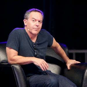 Comedian Greg Gutfeld sits on stage in a chair wearing a microphone headset.