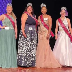 Pageant contenders line up on stage in ballgowns, crowns, and sashes