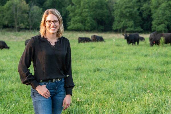 Photo of young woman standing in a green field with black angus cows