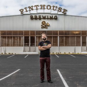 A man with this arms crossed standing in a parking lot in front a building labeled "PINTHOUSE BREWING"