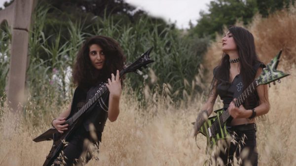 In this still image from the film "Sirens," two women with dark hair and black clothes stand in a field, playing electric guitars.