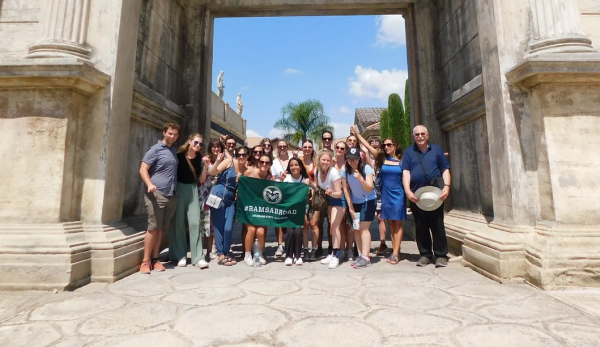 Group of students and professors standing together between white stone pillars holding a dark green sign that reads "#RAMSABROAD Colorado State University"