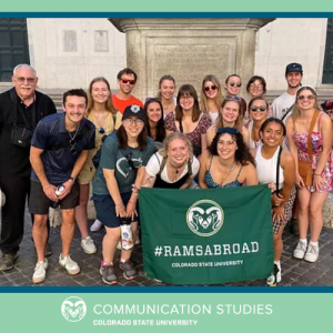 photograph of class standing outside on cobblestone street holding a green sign that reads "#RAMSABROAD Colorado State University" with a cartoon globe and airplane image on top