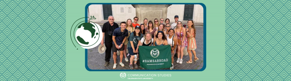 photograph of class standing outside on cobblestone street holding a green sign that reads "#RAMSABROAD Colorado State University" with a cartoon globe and airplane image on top