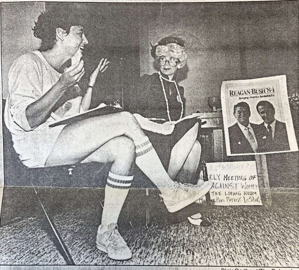 Black-and-white copy of newsprint photograph of two women in chairs talking, one in casual wear with long athletic socks and the other in a costume with a gray wig and hat, a pearl necklace, and a dress. Behind them is a poster that says "REAGAN-BUSH '84: Bringing America" and then someone has written "BACKWARDS" on top of the last word. There is also a handwritten sign on the floor behind the woman in the costume advertising the group's meeting.