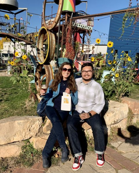 Associate Professor Julia Khrebtan-Hörhager and Joel Delgado sitting together outside the Lyric with metal art and blooming sunflowers behind them. Khrebtan-Hörhager is holding a cocktail and a shopping bag
