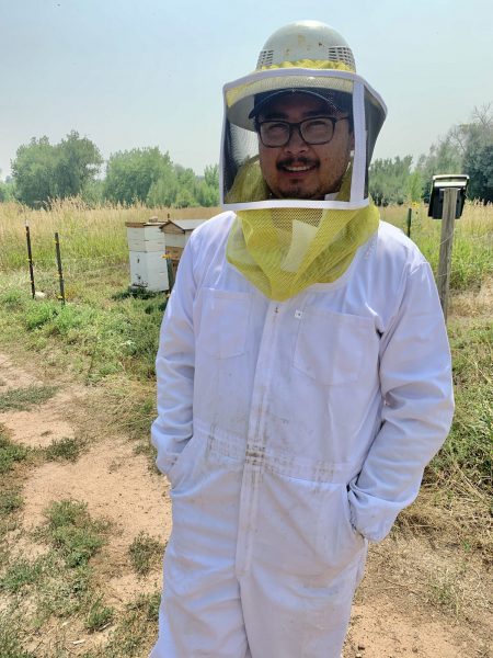Photo of Joel Delgado in a white beekeeper's suit and headnet out in a field