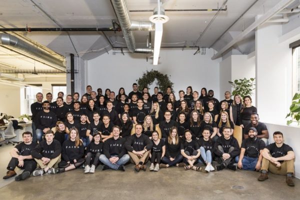 Photo inside an open office space of several dozen people standing and sitting together in a group, all wearing a black T-shirt that says "FAIRE"