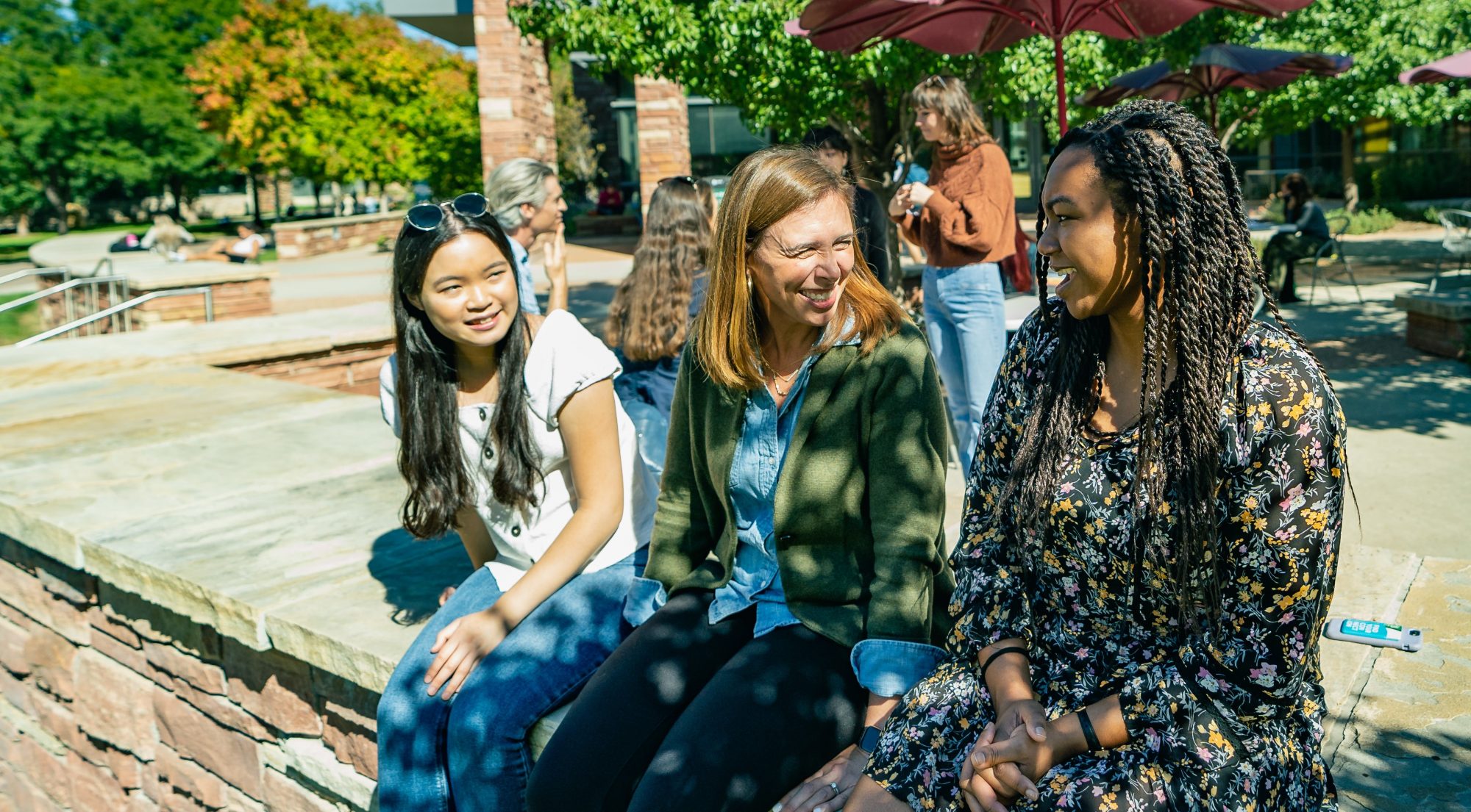 Two smiling female graduate students of color sit on a retaining wall outside talking with a smiling White female professor
