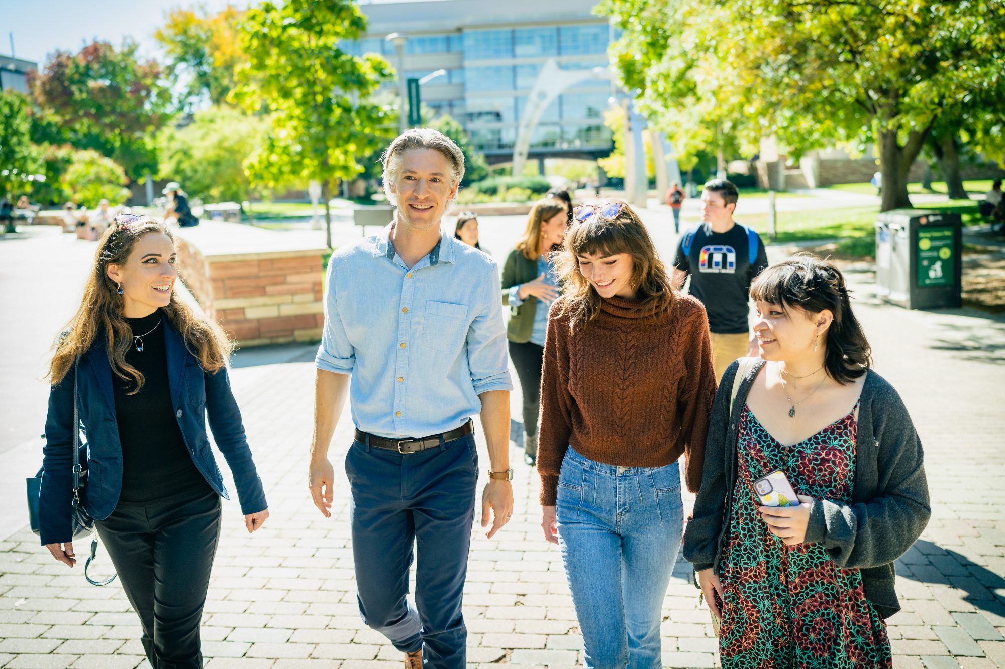 Two professors, one woman and one man, and two undergraduate students, both women, walk and talk while smiling together outside. Brick sidewalk and green leafy trees are in the background, as well as some other students