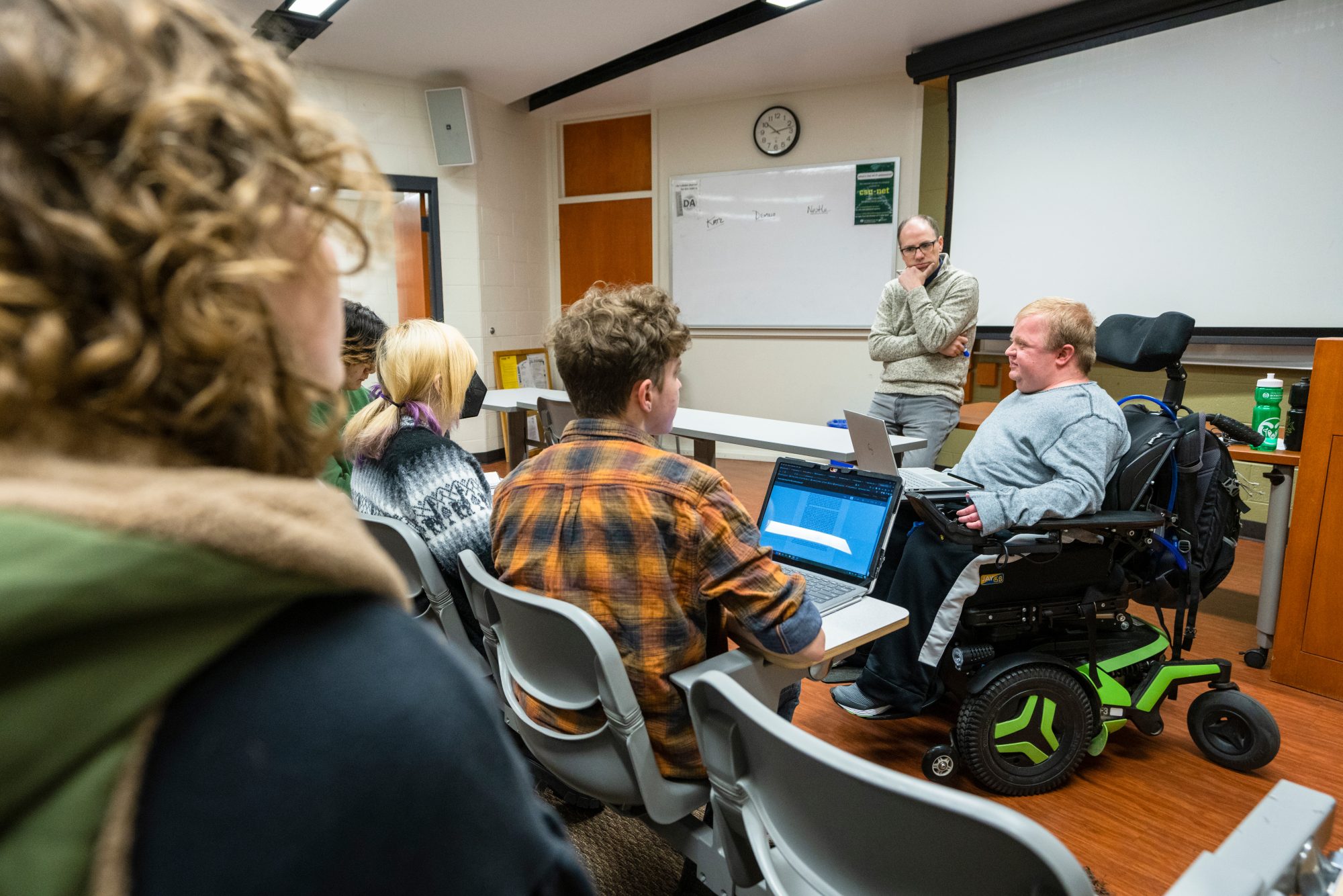 A classroom of college students listens to a presentation by a man in a wheelchair