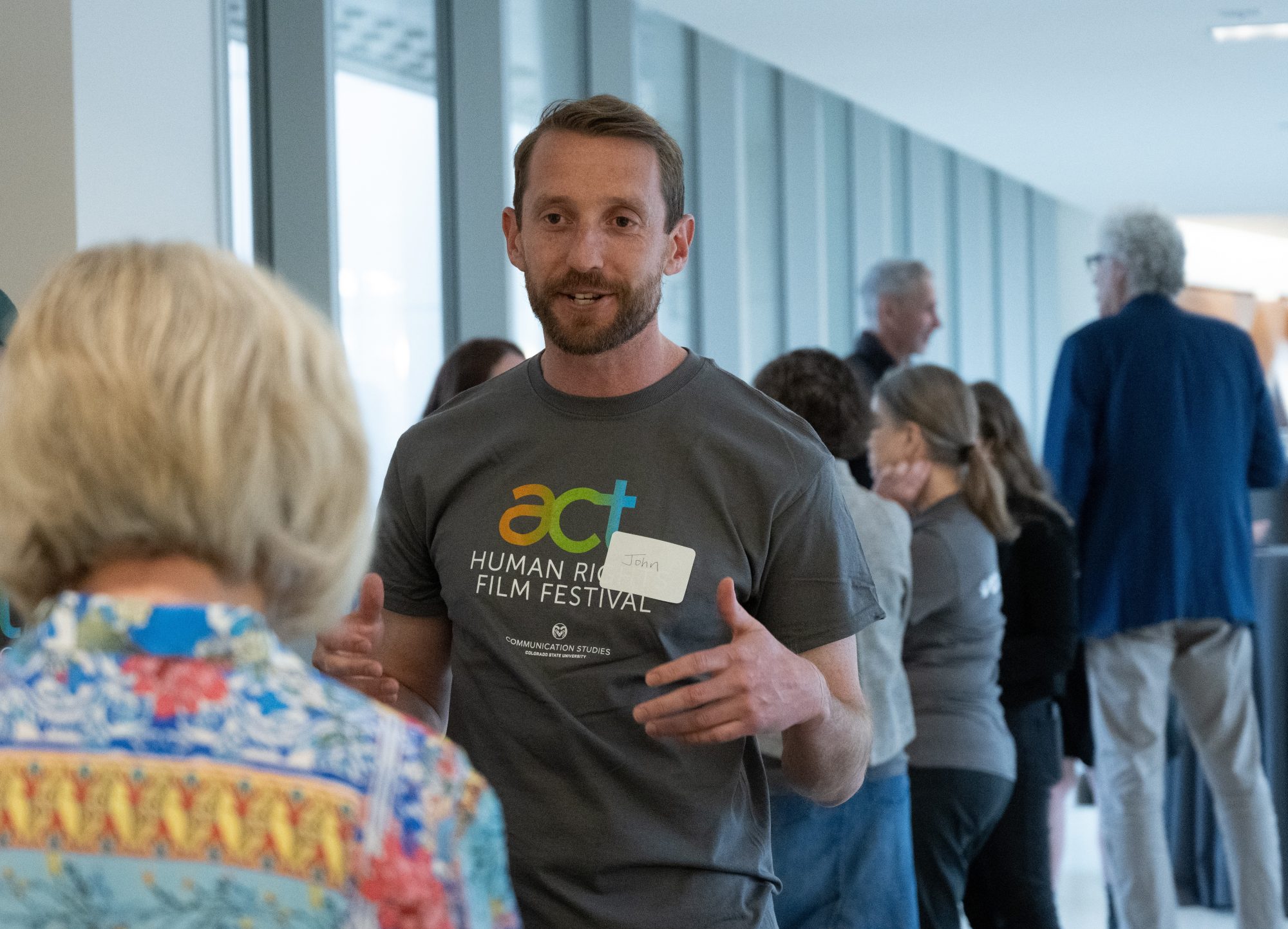 A bearded man wearing an ACT Human Rights Film Festival T-shirt and a handwritten nametag that says "John" speaks with a woman. Behind him folks are gathered in a hallway, chatting.
