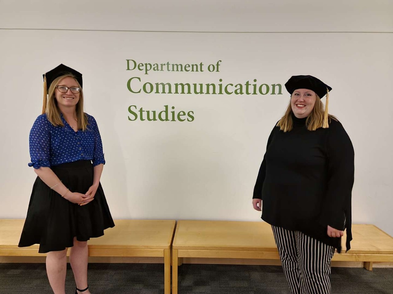 Two PhD graduates stand wearing their graduation caps before a sign that says "Department of Communication Studies"