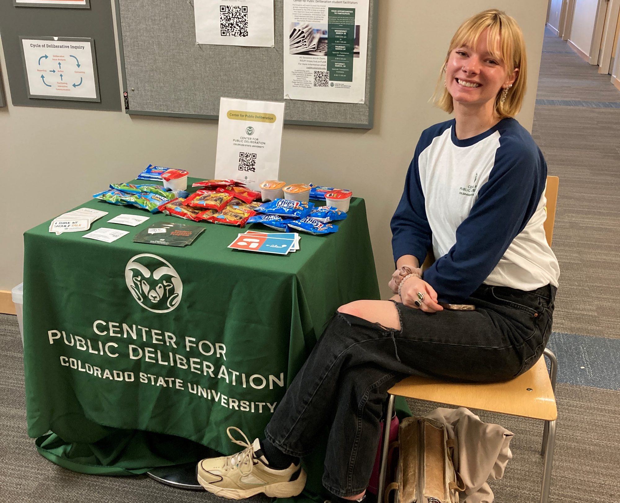 A college student smiles seated next to a small table in a hallway with a tablecloth that says "Center for Public Deliberation"