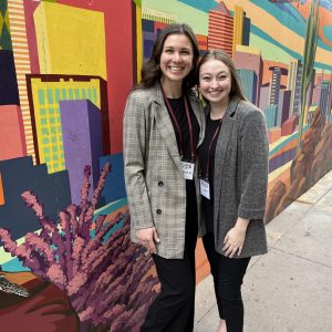 Two women in professional attire smiling in front of a colorful wall mural
