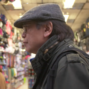 Photograph from the side of photographer Corky Lee, an older Asian man wearing a gray newsboy hat and wearing glasses and a coat standing inside a store
