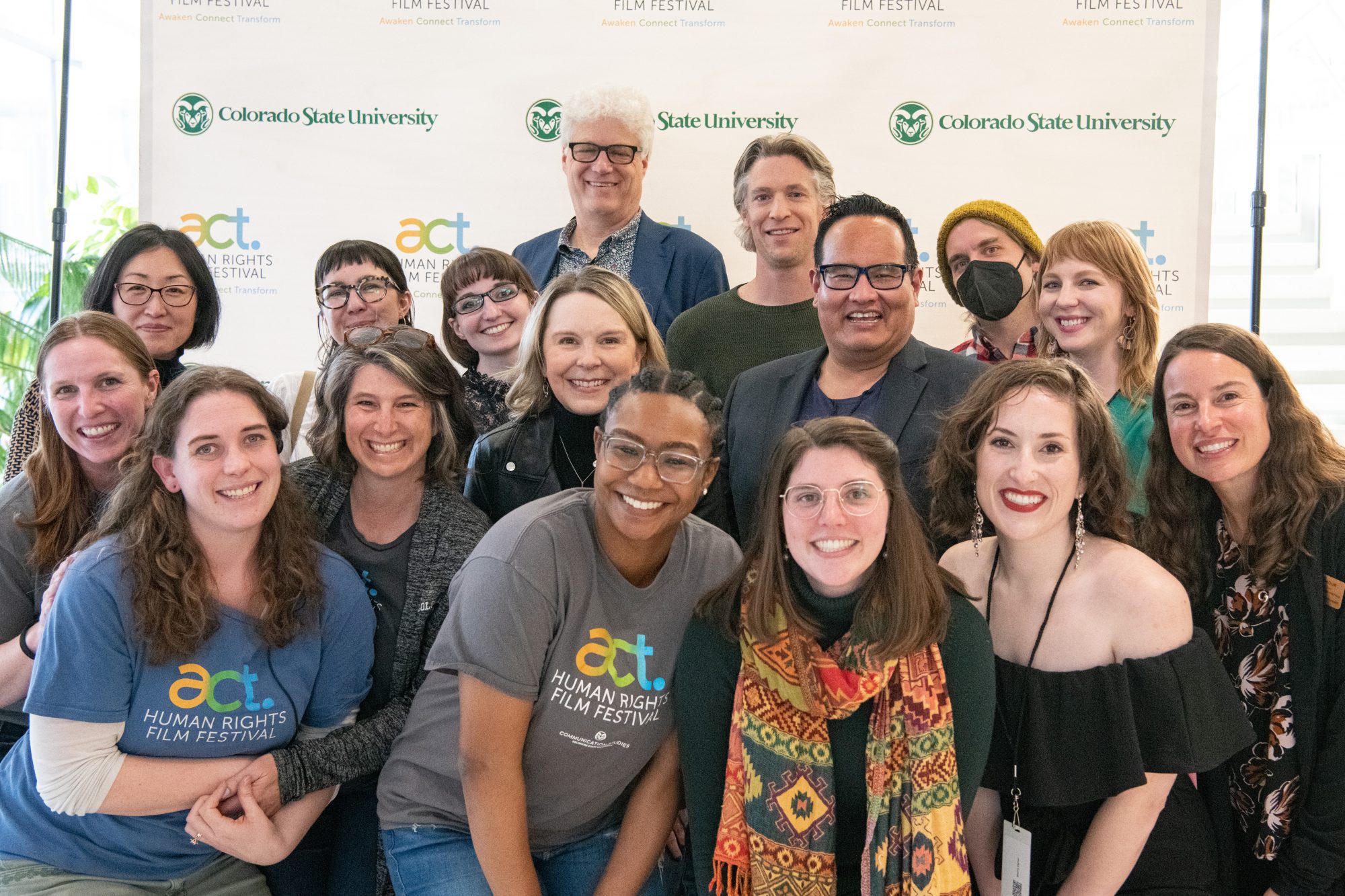 A group of 16 people stands together smiling in front of a white background that reads "Colorado State University, ACT Human Rights Film Festival: Awaken, Connect, Transform"