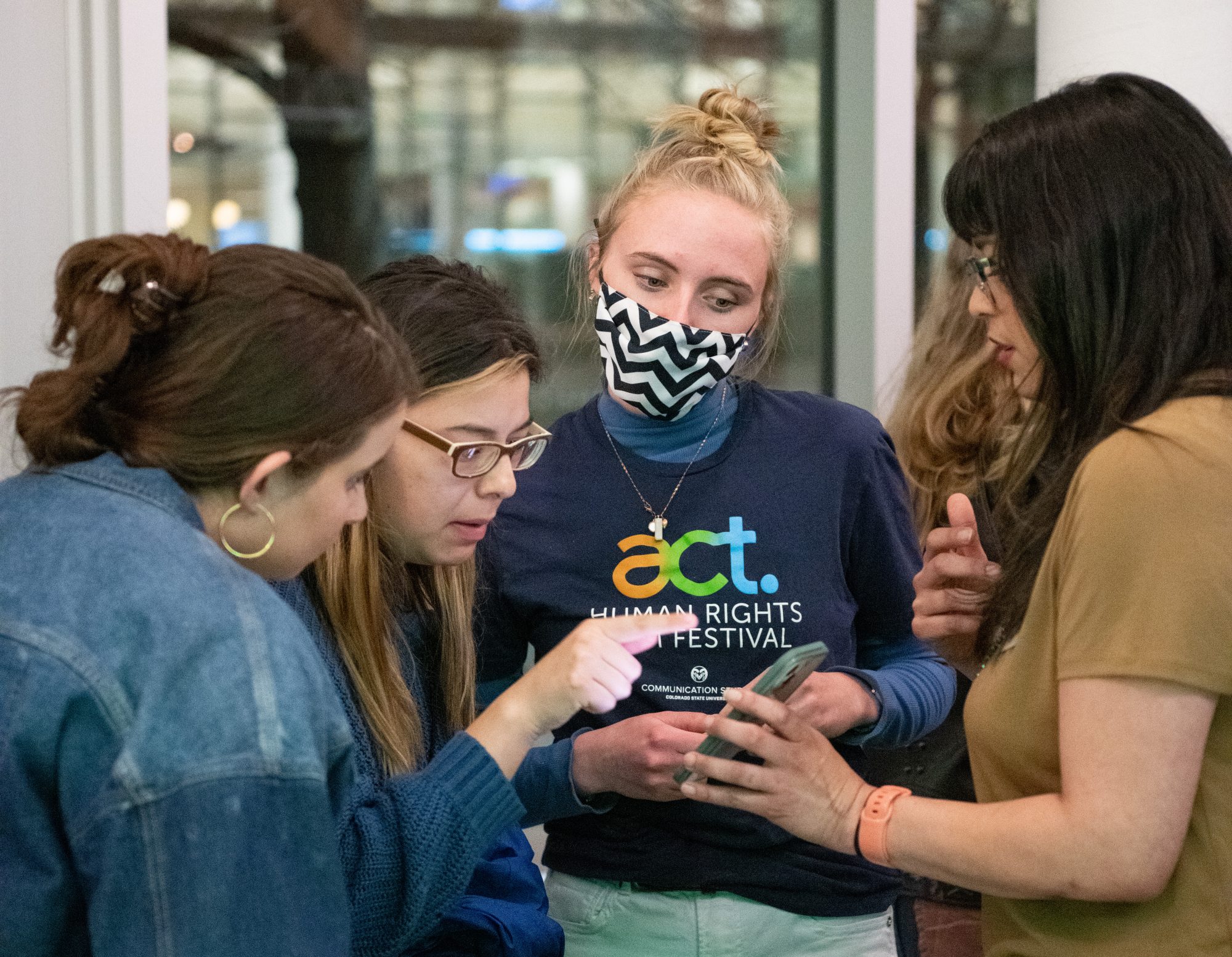A group of 4 students stands together, looking at a smartphone. The student holding the phone is wearing a facemask and a T-shirt that reads "ACT Human Rights Film Festival"