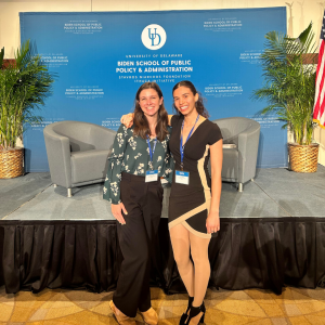 Two college students, Olivia Birg and Abby Brier, stand and smile with their arms around one another before a stage with a blue backdrop that reads "University of Delaware Biden School of Public Policy & Administration Stavros Niarchos Foundation Ithaca Initiative"