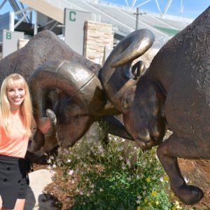 Photo of a young woman with long, blonde hair dressed professionally and smiling next to a large, bronze statue of two rams butting heads outside the football stadium on CSU's campus