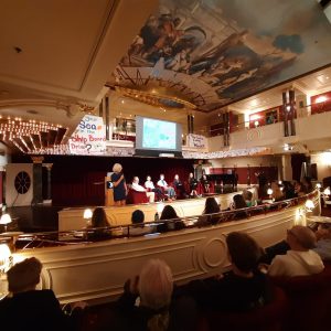 Photo of a room on a ship warmly lit with an audience watching a panel discussion on a stage. A painted mural is on the ceiling.