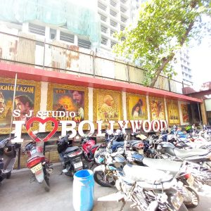 Photo of a building with a yellow facade and a big sculpture with white letters reading "I Heart Bollywood" before a parking lot full of parked motorbikes