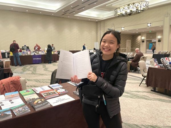 Madison Barnes-Nelson smiles while holding open a book to the page where her chapter in it begins. Behind her is a conference room with a book fair, containing tables with books on display atop them.