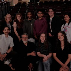 A diverse group of nine college students and one professor pose together in a theater, smiling