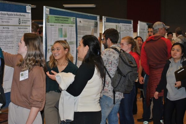 Photo of many college students and other adults standing in a room in front of several research posters on display, looking at and talking about them with each other