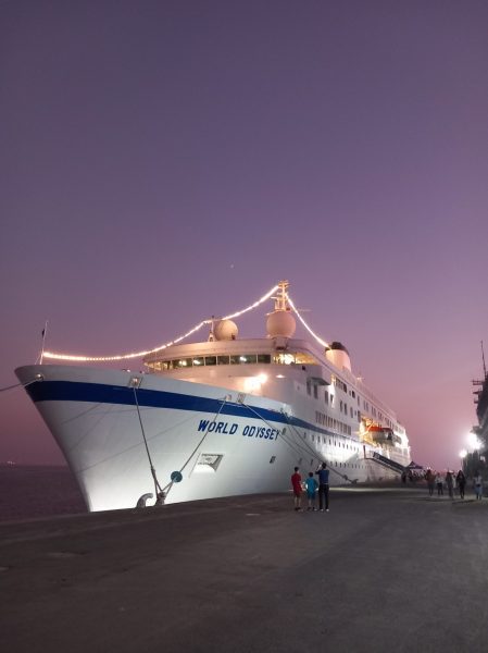 Photo of a large white ship, lit up at dusk with a purple sky behind it, docked. There is a blue stripe around the ship with blue text "WORLD ODYSSEY" on its side.