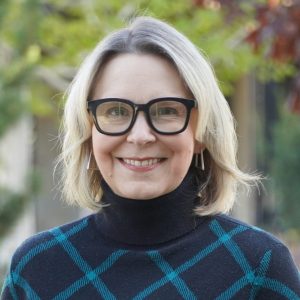 Headshot of Dr. Karrin Vasby Anderson wearing a blue checkered sweater outside with blurred trees in the background