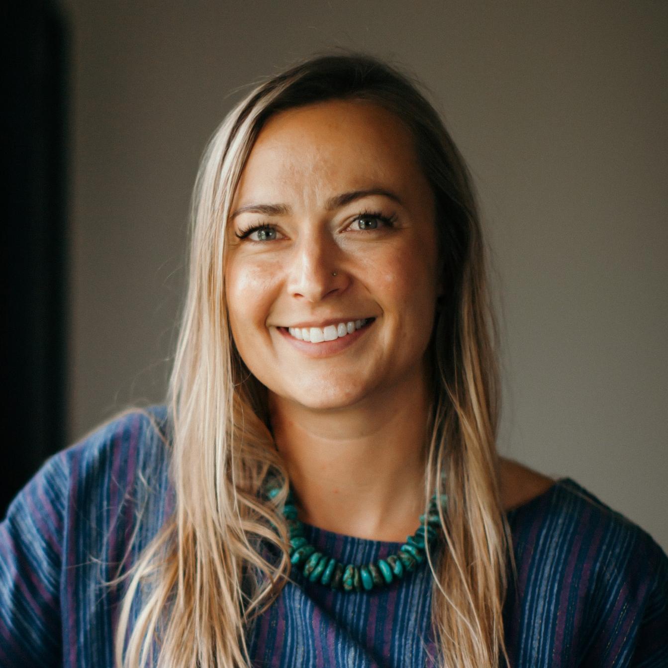 Headshot of Emily Amedee with a blue sweater and turquoise necklace against a grey background