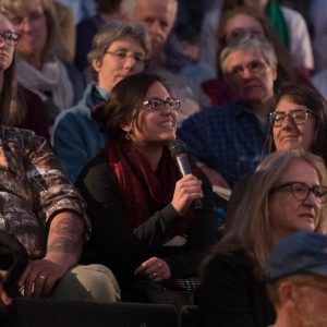 an audience of people sitting in theater seats. One is holding a microphone to ask a question