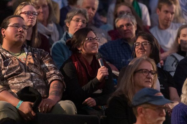 an audience of people sitting in theater seats. One is holding a microphone to ask a question