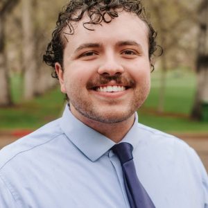 Headshot of John outside with a dress shirt tie and curly hair