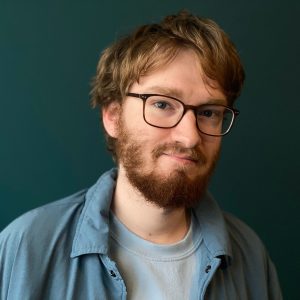 Headshot of Evan Bode with a blue button up and a blue background