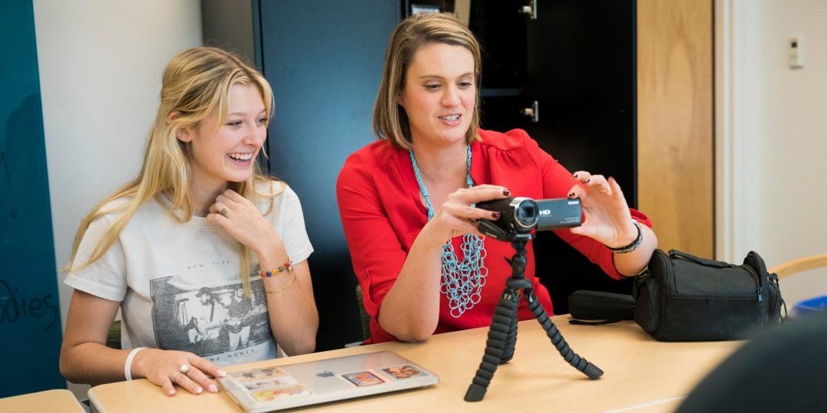 A professor and college student sit at a table together, both smiling. The professor is manipulating a video camera on a tripod on the table. A closed laptop sits before the student.