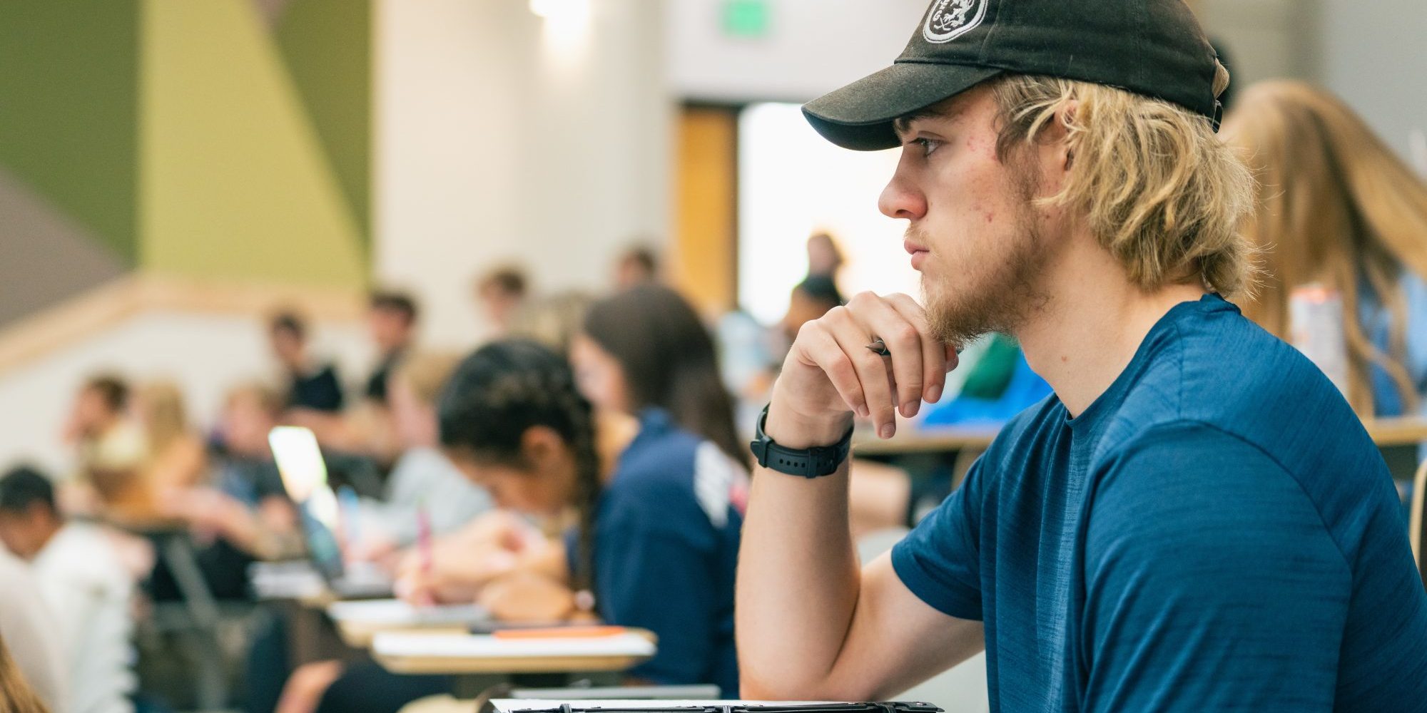 Photo of a young man with blond hair and a beard wearing a ball cap paying attention in a classroom