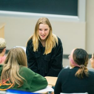 Female college student stands in a classroom, speaking to her fellow students