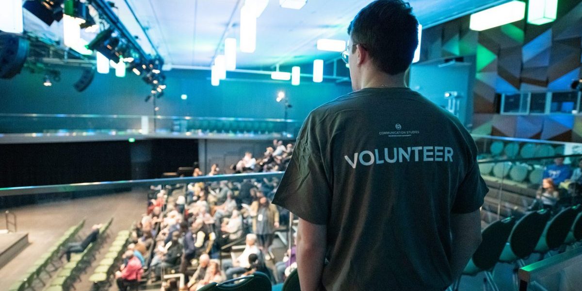 An adult facing away from the camera wears a T-shirt that says "VOLUNTEER" on the back, looking out over a full theater.