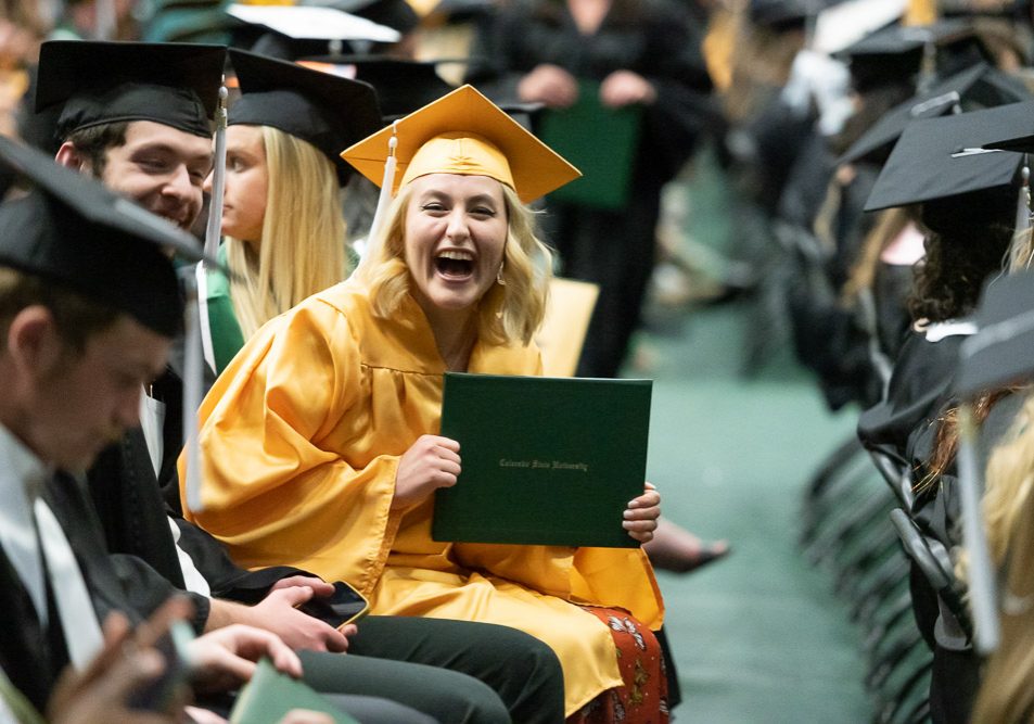 A grinning college graduate in a yellow cap and gown holds her Colorado State University diploma cover, surrounded by other graduates in caps and gowns